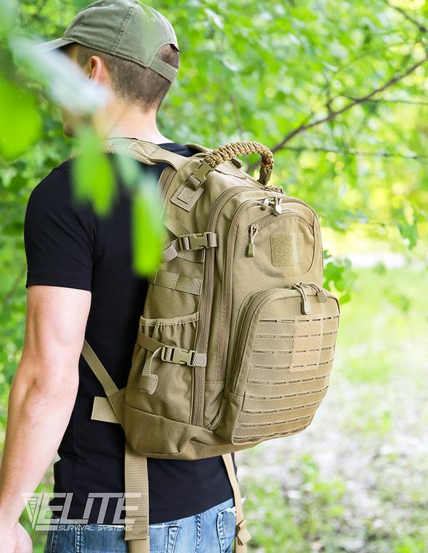 Man wearing a black t-shirt and cap, carrying a tan Elite Survival Systems PULSE 24-Hour Backpack in a lush green forest.