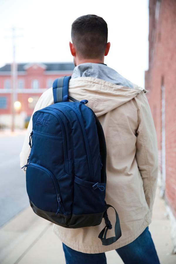 Man walking down a city street, viewed from behind, wearing a beige jacket and carrying an Elite Survival Systems Echo EDC Backpacks 7721-TR.