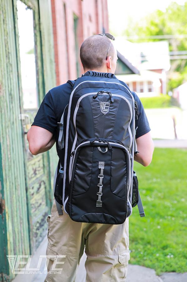 A man with a shaved head wearing an Elite Survival Systems STEALTH Covert Operations Backpack stands facing away on a sidewalk near a weathered green door.