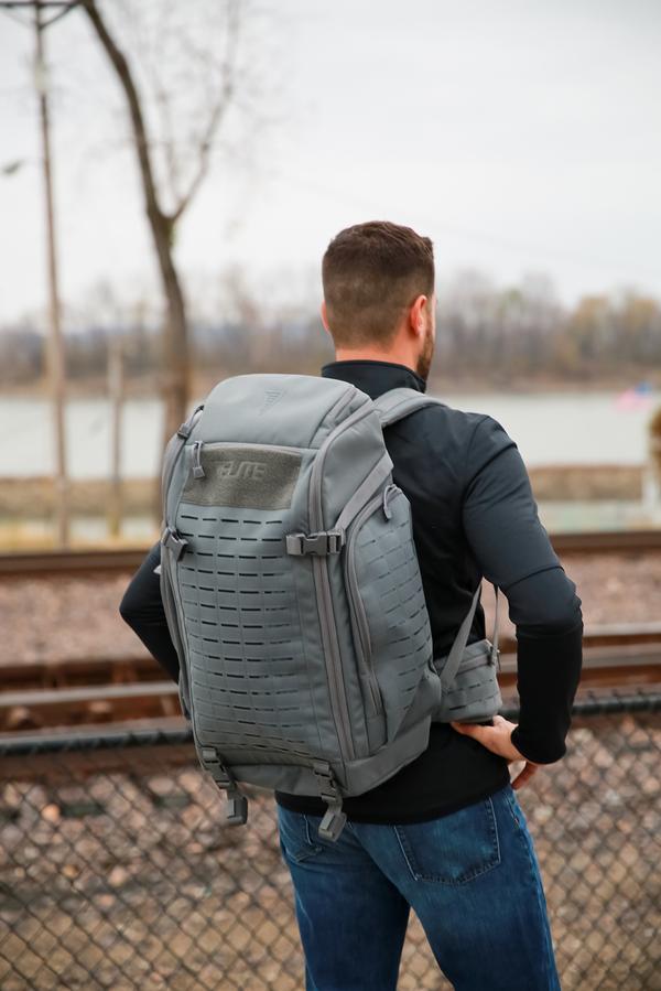 Man wearing a Elite Survival Systems Tenacity-72 Three Day Support/Specialization backpack stands facing away, by railroad tracks with bare trees in the background.