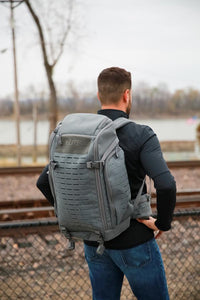 Thumbnail for Man wearing a Elite Survival Systems Tenacity-72 Three Day Support/Specialization backpack stands facing away, by railroad tracks with bare trees in the background.