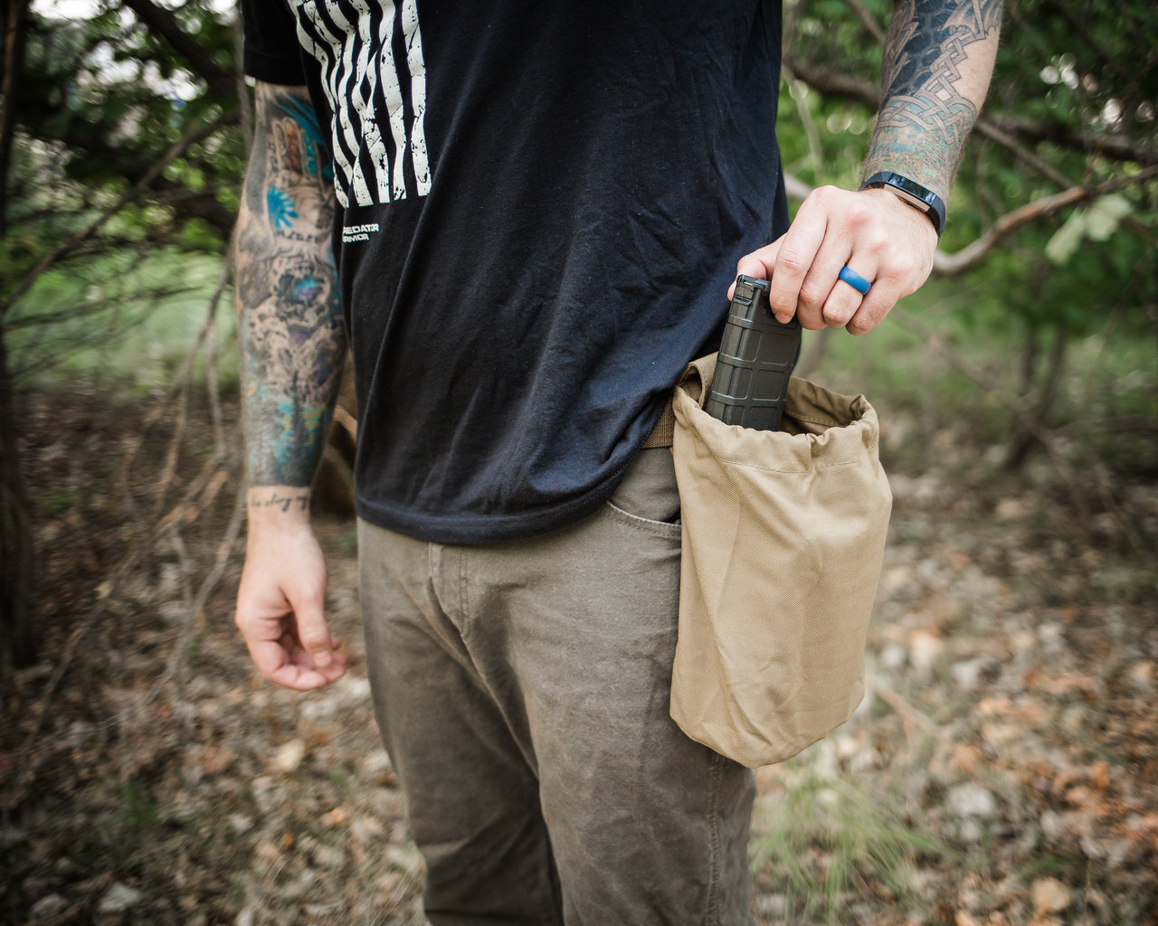 Man with tattooed arms holding a black water bottle in a Predator Armor Dump Pouch attached to his belt, standing in a wooded area.