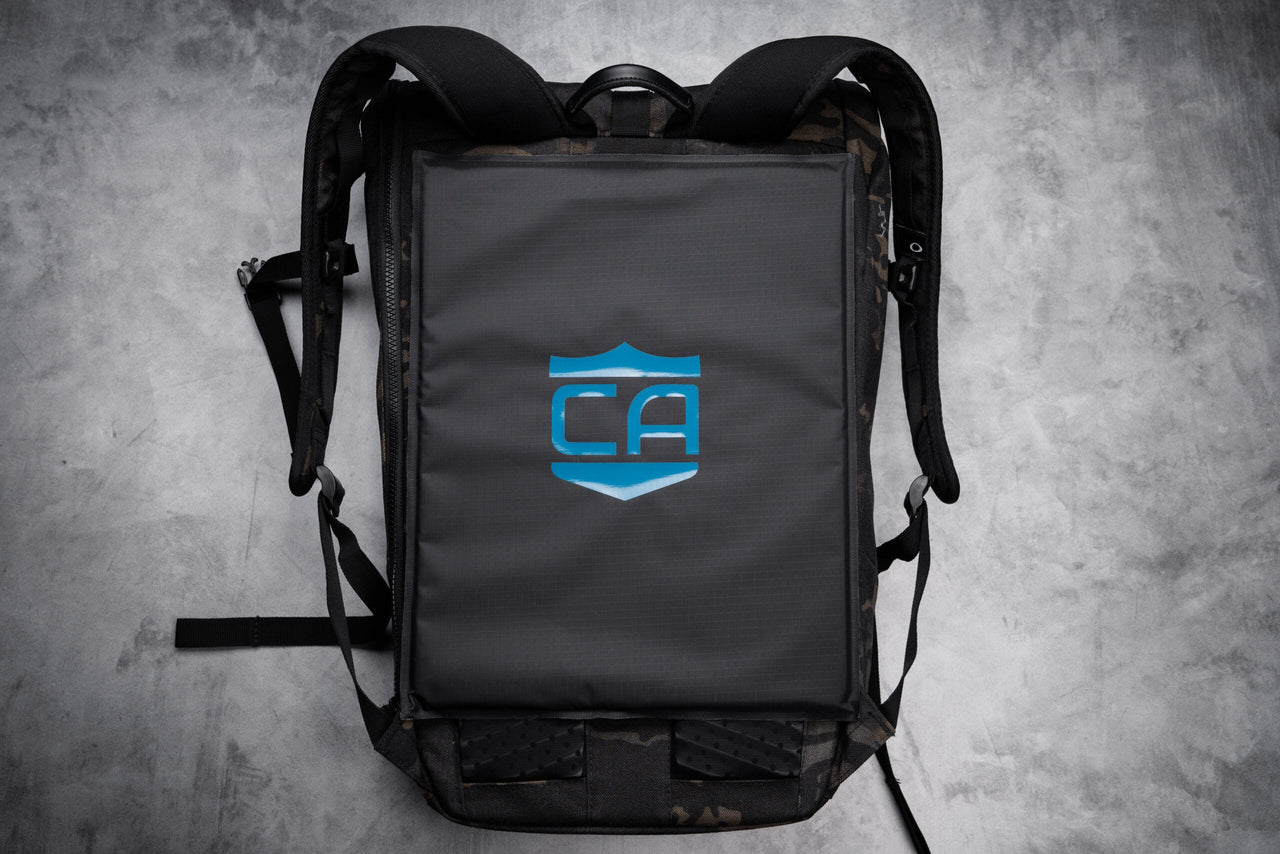 A black backpack lies flat on a gray surface, featuring a blue "CA" shield logo and designed to fit the Caliber Armor CaliberX IIIA Soft Body Armor Rectangle Panel for enhanced protection.