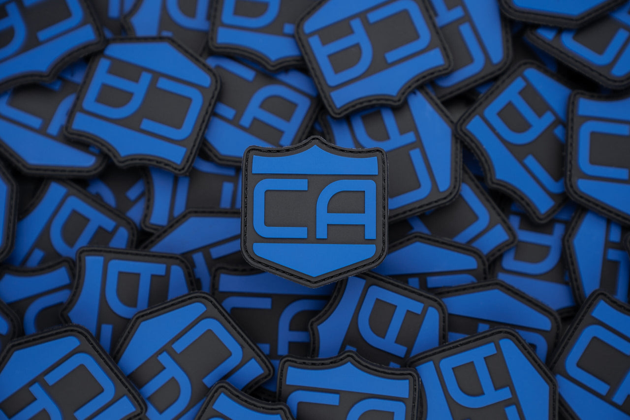 A stack of blue and black shield-shaped Caliber PVC Morale Patches by Caliber Armor, each with "CA" at the center—an eye-catching Guardian Shield design ideal for any tactical gear collection.