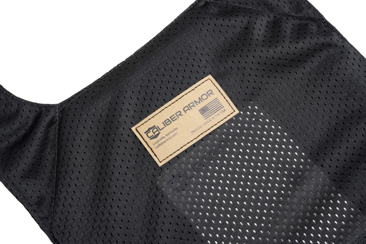 Close-up of the Gray Man Concealable Carrier by Caliber Armor, made from black mesh fabric with a tan Caliber Armor label and an American flag patch.