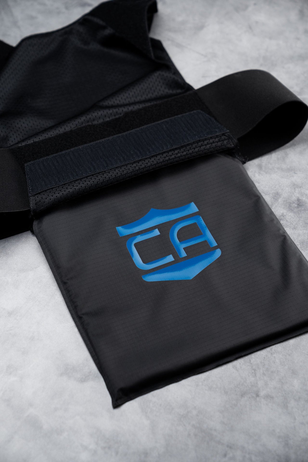 The Caliber Armor CaliberX IIIA Gray Man Concealable Package is a black padded pouch with a blue geometric logo, designed for discreet carry or soft armor panels, attached to a strap and Velcro on a light gray textured surface.