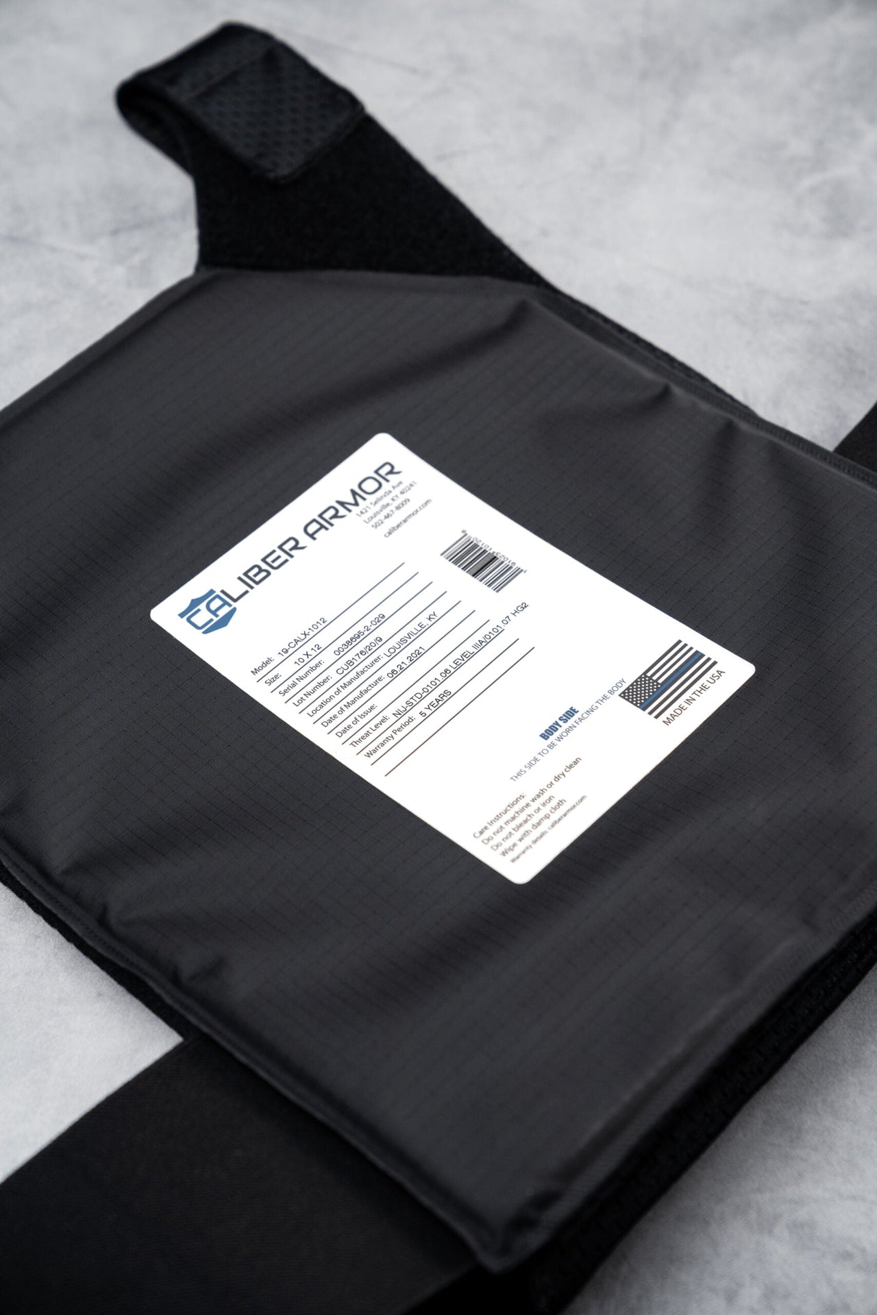 A close-up of a Caliber Armor CaliberX IIIA Gray Man Concealable Package body armor plate with a white product info label and barcode, shown on a light gray surface.