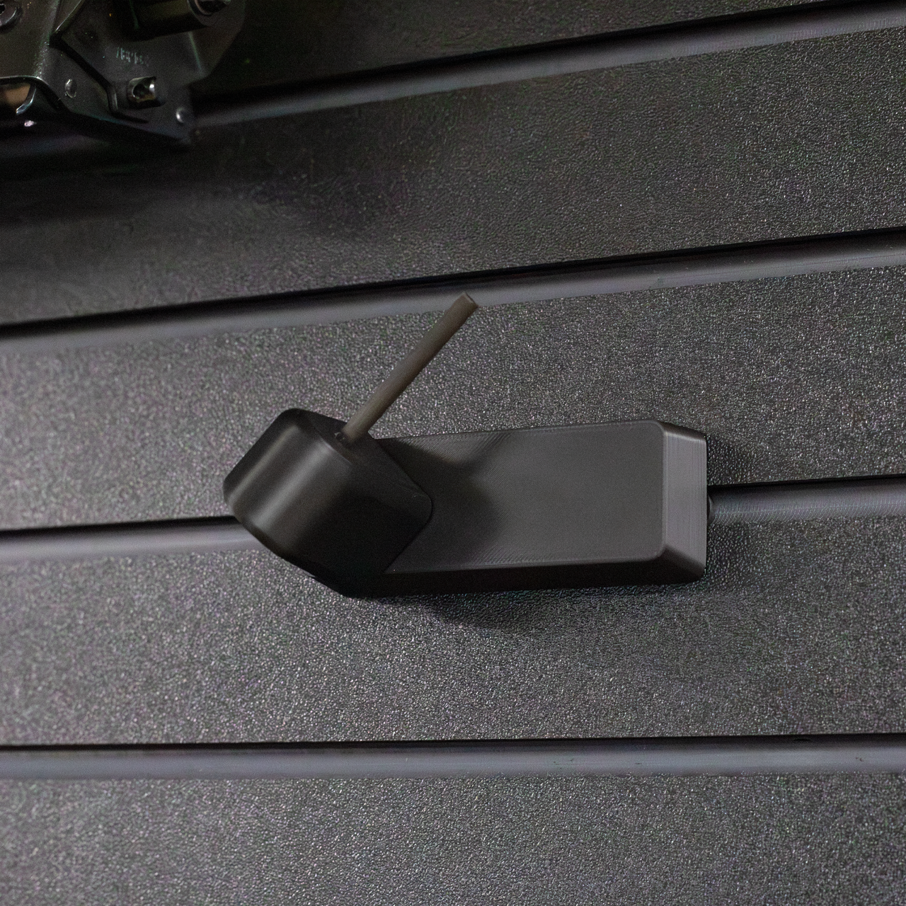 Black plastic barrel mounted pistol hanger that has a carbon fiber rod, shown mounted to our Assault Wall tactical slat wall panel