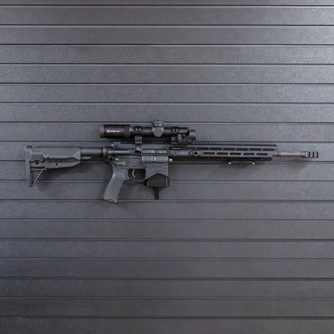 Right facing AR-15 rifle mounted by its magazine well on a black Assault wall slat wall panel