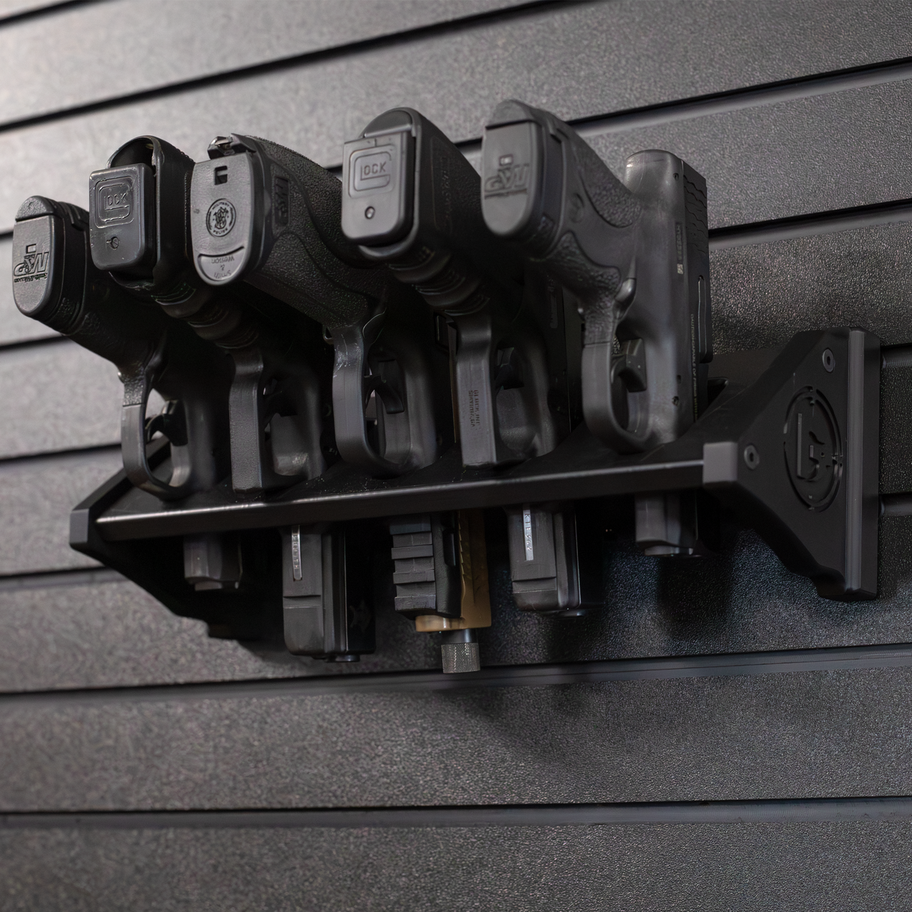 Black plastic five pistol storage mount loaded with smith and weston and glock pistols , all five pistols are resting in equally spaced slots, mount is hanging from black tactical slat wall