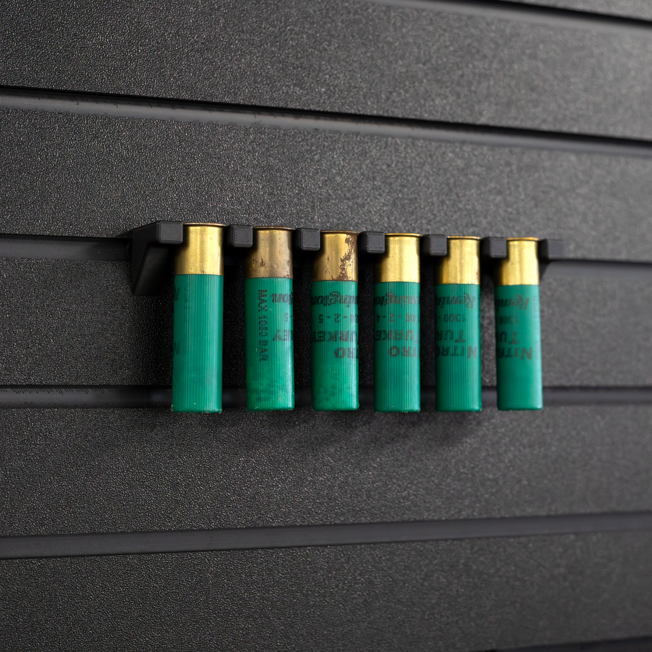 Six green 12 gauge shotgun shells hanging from a black mount that is attached to an Assault Wall tactical slat wall panel