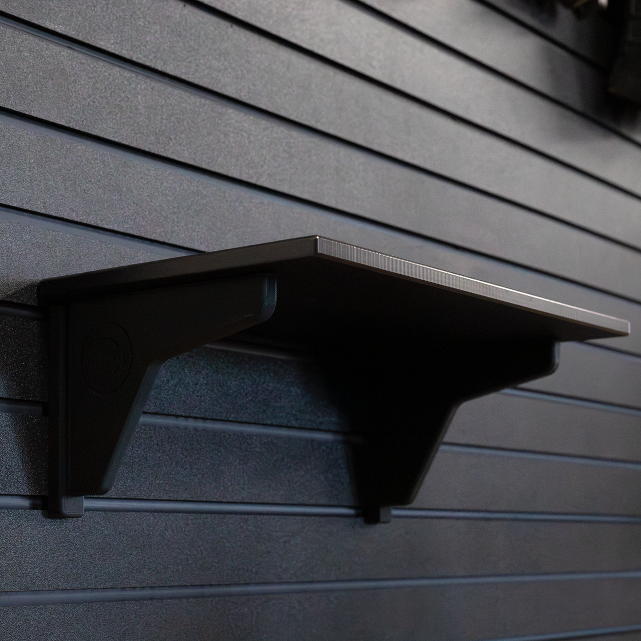 15" black plastic shelf with two support backets mounted on a black tactical slat wall