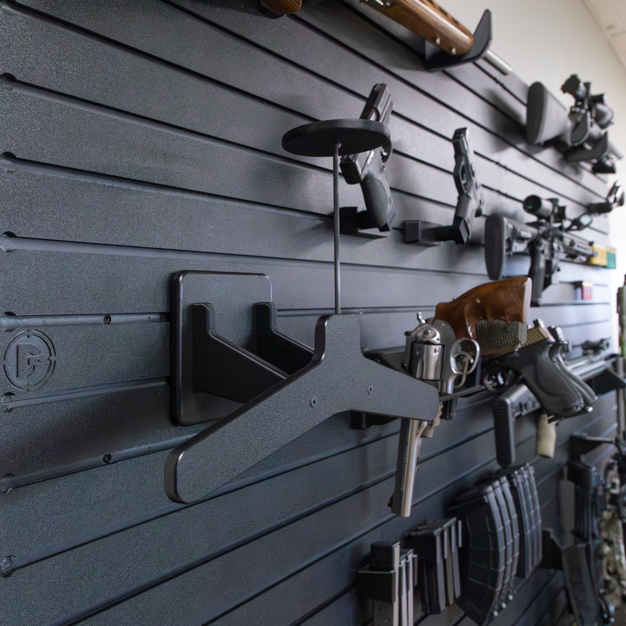 Empty black plastic plate carrier and helmet hanging system meant for heavy duty tactical gear mounted to Assault Wall slat wall with guns and accessories organized in the blurred background