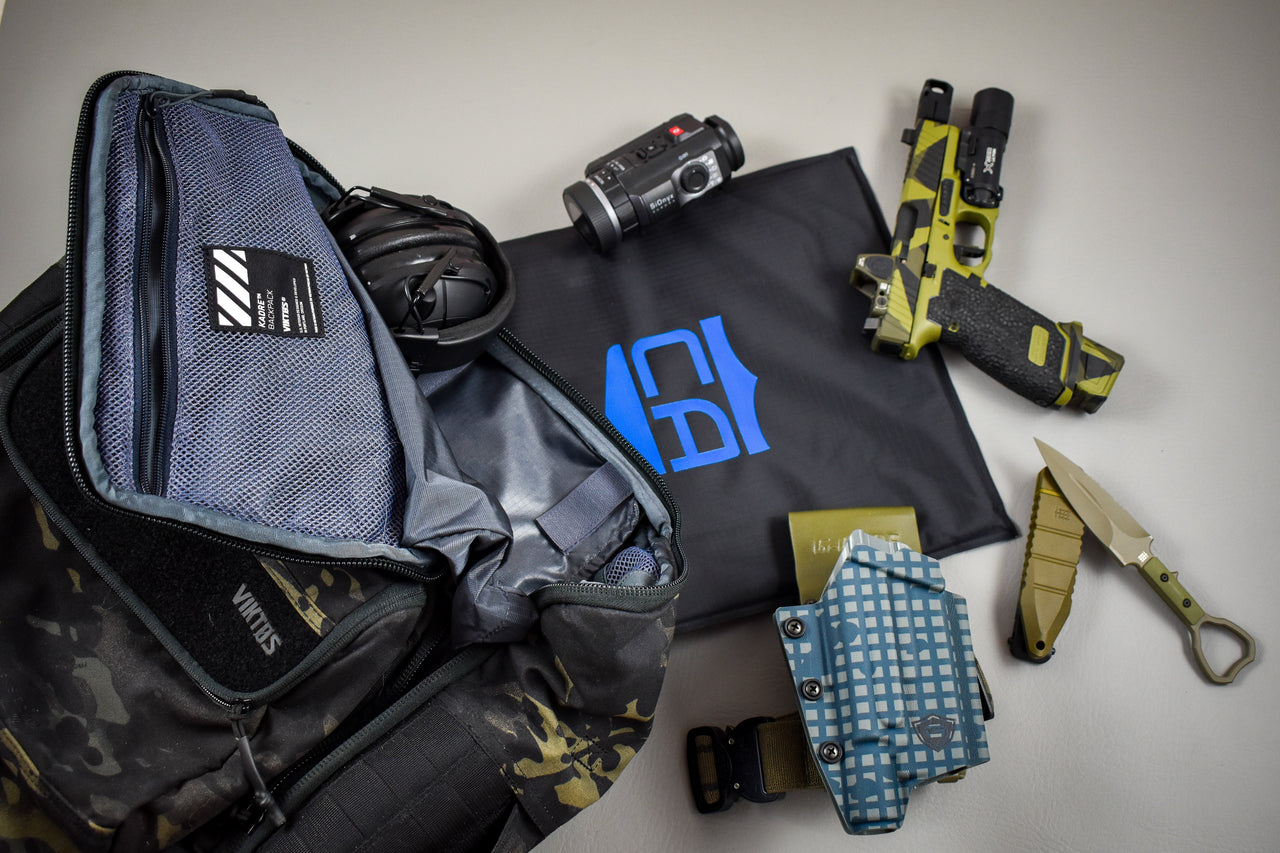 A 5.11 Tactical Backpack displays gear—headphones, camera, folding knife, holstered pistol, and a black mat with a blue shield logo—featuring the CaliberX IIIA Body Armor Insert from Caliber Armor on a grey surface.