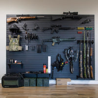 Thumbnail for Assault Wall systems, black plastic wall-mounted slat wall display of various firearms and tactical gear in a room with wooden flooring.