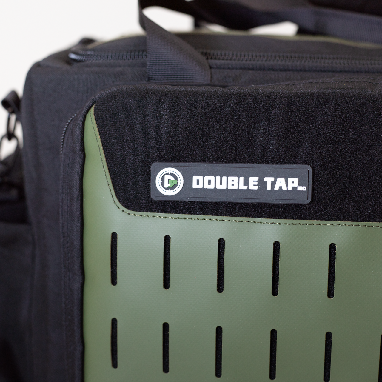 Close-up of a green and black bag adorned with the Double Tap Industries Logo Velcro Patch. The bag’s vertical slotted design on the green panel adds tactical flair, making it a stylish gear accessory.