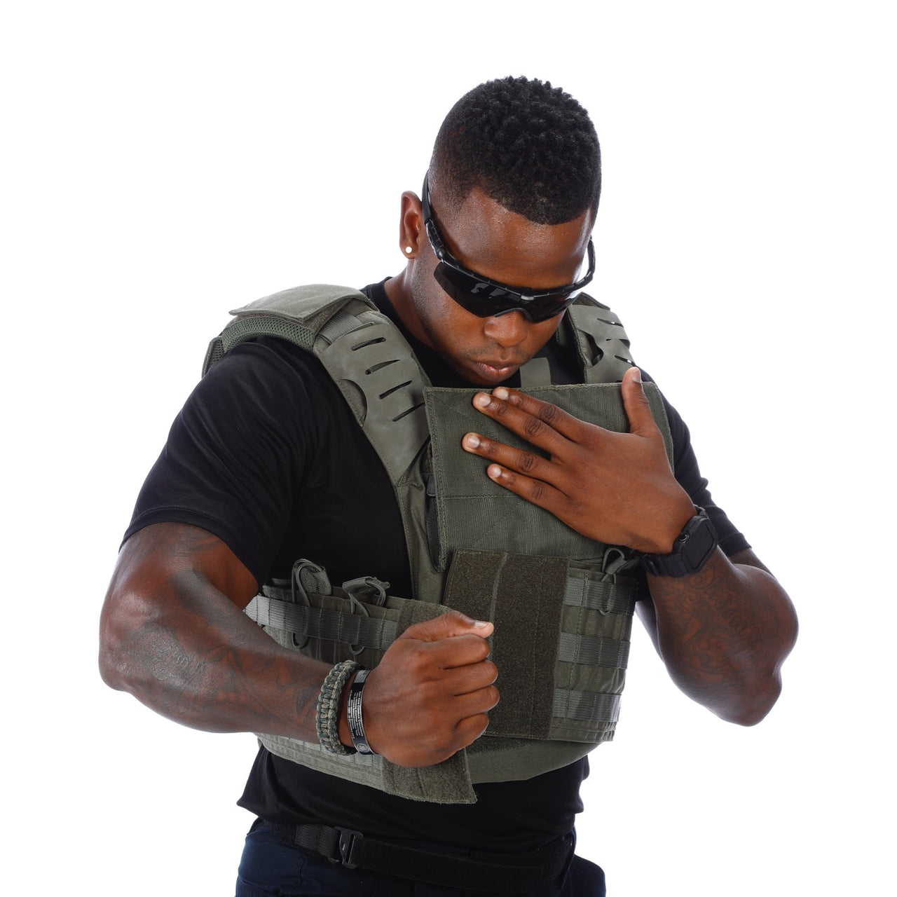 A man wearing a Body Armor Direct Victory Plate Carrier Deluxe Edition and sunglasses checks his watch while looking down, isolated on a white background.