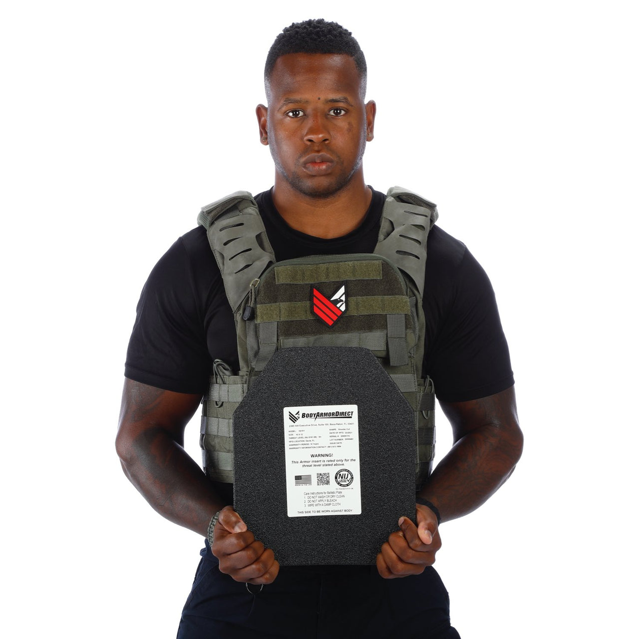 A man in a black t-shirt and Body Armor Direct Victory Plate Carrier Deluxe Edition holds a body armor plate facing forward against a white background.